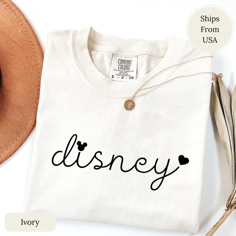 Puede incluir: Camiseta de color marfil con la palabra "disney" en escritura negra, con un icono de la cabeza de Mickey Mouse encima de la "d" y un coraz&oacute;n al final. La camiseta tiene cuello redondo y una etiqueta Comfort Colors.