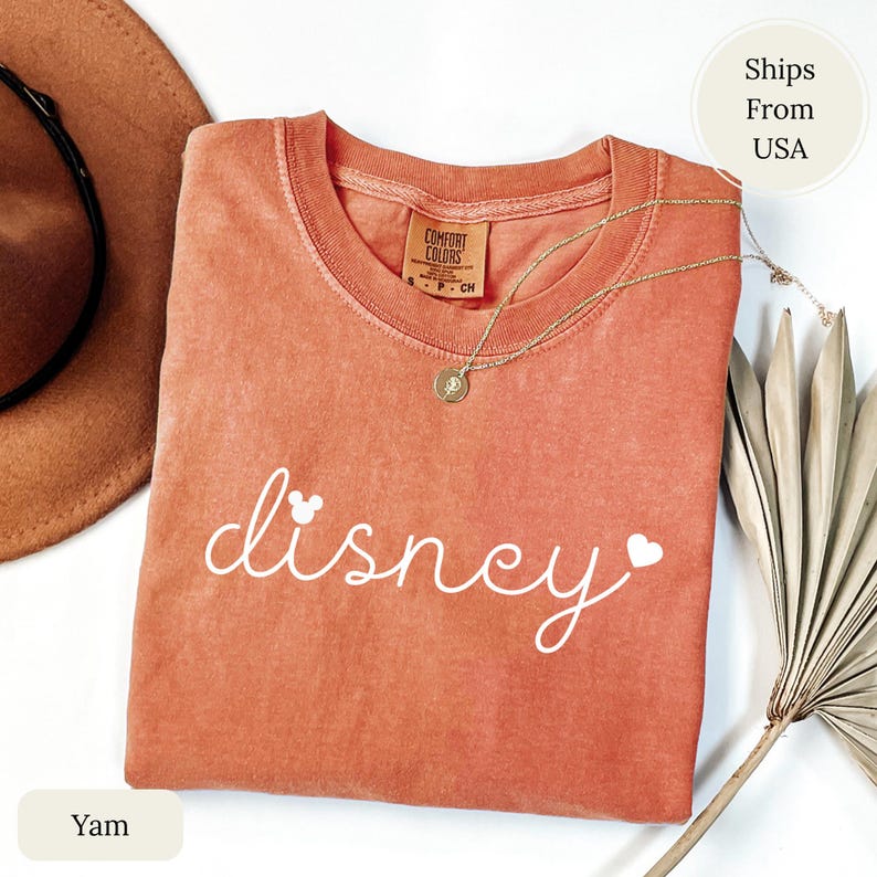 Puede incluir: Una camiseta Comfort Colors de color coral con la palabra "disney" en escritura blanca, con una cabeza de Mickey Mouse y un coraz&oacute;n. Un collar de oro con un colgante descansa sobre la camisa. Un sombrero marr&oacute;n y una hoja de palma seca tambi&eacute;n est&aacute;n en la imagen.