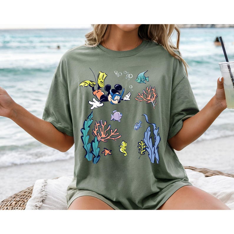 May include: Olive green t-shirt featuring a cartoon Mickey Mouse snorkeling underwater with colorful coral, fish, and bubbles. The design includes yellow flippers and a black snorkel mask. The shirt is a relaxed fit.