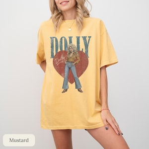 K&ouml;nnte beinhalten: Ein senfgelbes Kurzarm-T-Shirt mit einem gro&szlig;en roten Herz-Aufdruck auf der Brust. Im Herz befindet sich die Illustration einer blonden Person in einem beigen Hemd und blauen Jeans. Der Text DOLLY erscheint in gro&szlig;en blauen Buchstaben dar&uuml;ber.