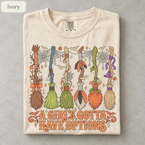 May include: An ivory t-shirt featuring a graphic of six distinct witch brooms with colorful bristles, spiderwebs, bats, and small pumpkins. Below the brooms, the text reads A GIRL'S GOTTA HAVE OPTIONS in orange lettering with decorative flourishes.