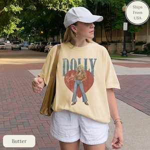 K&ouml;nnte beinhalten: Ein buttergelbes Kurzarm-T-Shirt mit einer Grafik einer Frau in beigem Hemd und blauer Jeans, die in einem gro&szlig;en roten Herzen steht. Das Wort DOLLY ist in gro&szlig;en blauen Buchstaben &uuml;ber dem Herzen gedruckt, mit dem Namen Dolly in Schreibschrift.