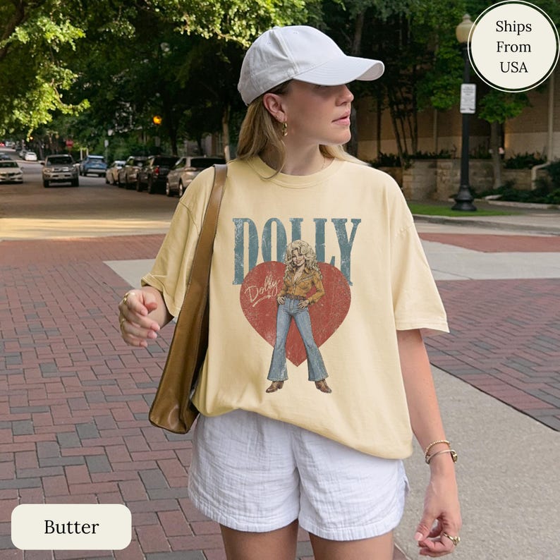 K&ouml;nnte beinhalten: Ein buttergelbes Kurzarm-T-Shirt mit einer Grafik einer Frau in beigem Hemd und blauer Jeans, die in einem gro&szlig;en roten Herzen steht. Das Wort DOLLY ist in gro&szlig;en blauen Buchstaben &uuml;ber dem Herzen gedruckt, mit dem Namen Dolly in Schreibschrift.