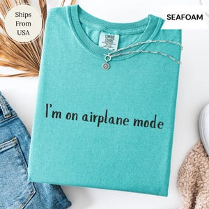 May include: Seafoam green t-shirt with the phrase "I'm on airplane mode" printed in black. A silver necklace with a sun pendant rests on the shirt. The shirt is folded with a pair of blue jeans.