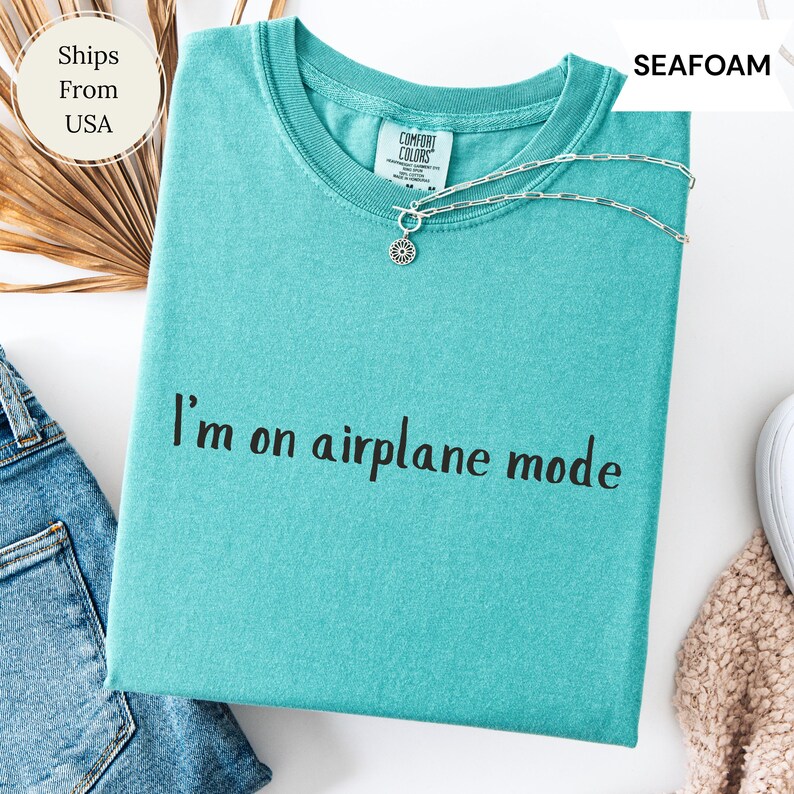 May include: Seafoam green t-shirt with the phrase "I'm on airplane mode" printed in black. A silver necklace with a sun pendant rests on the shirt. The shirt is folded with a pair of blue jeans.