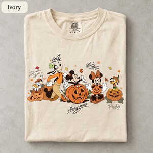 May include: An ivory t-shirt featuring a graphic of Disney characters including Donald Duck, Goofy, Mickey Mouse, Minnie Mouse, and Pluto interacting with jack-o'-lanterns. The design includes handwritten-style names and autumn leaf accents.