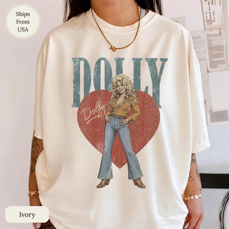 K&ouml;nnte beinhalten: Ein elfenbeinfarbenes Kurzarm-T-Shirt mit einem gro&szlig;en roten Herz und dem Wort DOLLY in blauen Blockbuchstaben dar&uuml;ber. Im Herz befindet sich die Illustration einer Person in einem beigen Hemd und einer blauen Schlaghose mit dem Namen Dolly.