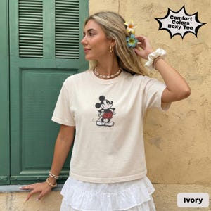 May include: Ivory-coloured boxy tee featuring a classic Mickey Mouse graphic. The shirt has short sleeves and a round neckline. The text "Comfort Colors Boxy Tee" is in a starburst shape. The shirt is paired with a white skirt.