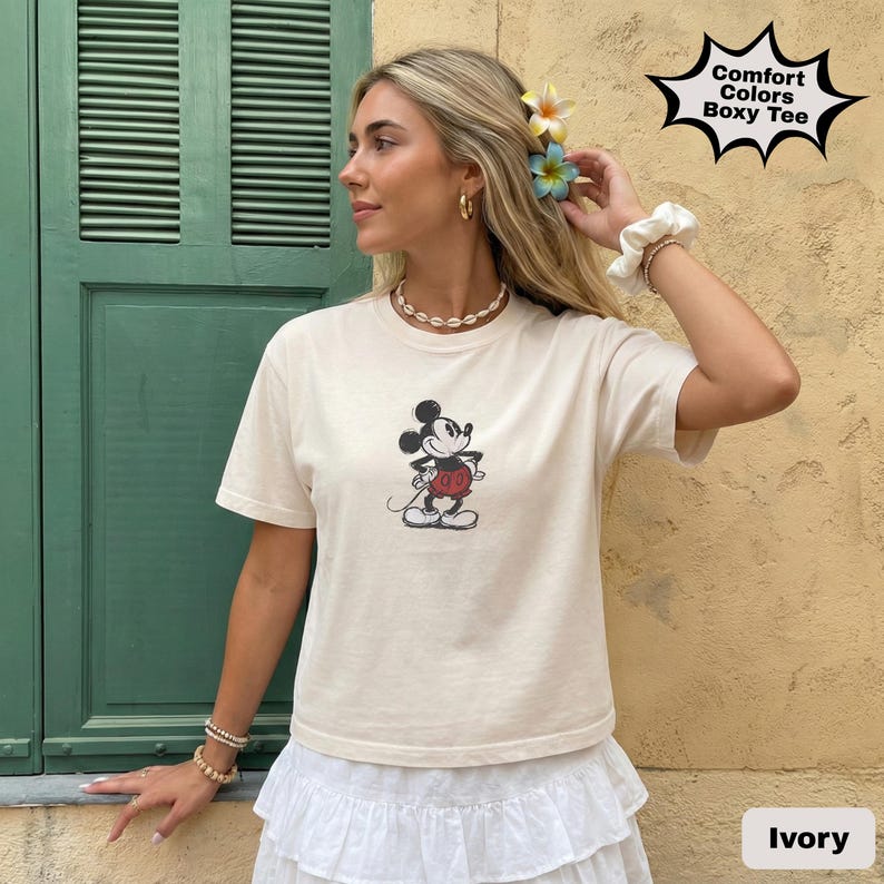 May include: Ivory-coloured boxy tee featuring a classic Mickey Mouse graphic. The shirt has short sleeves and a round neckline. The text "Comfort Colors Boxy Tee" is in a starburst shape. The shirt is paired with a white skirt.