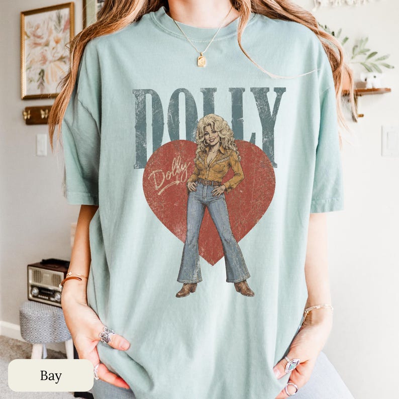 K&ouml;nnte beinhalten: Ein hellgr&uuml;nes Kurzarm-T-Shirt mit einem gro&szlig;en roten Herzmotiv in Used-Optik. In der Mitte befindet sich die Abbildung einer blonden Person in einem beigen Hemd und blauen Schlaghosen. Der Schriftzug DOLLY steht in gro&szlig;en Buchstaben dar&uuml;ber.