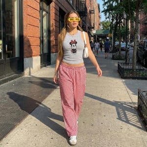 May include: A person wearing a grey vest top with a Minnie Mouse graphic, yellow sunglasses, and red and white gingham trousers. They are carrying a white bag and walking on a pavement in front of a brick building.