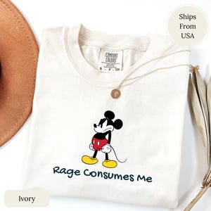 May include: Ivory-colored t-shirt featuring a cartoon Mickey Mouse graphic with the text "Rage Consumes Me." The graphic shows Mickey in his classic pose, with black ears, a red torso, yellow shoes, and white gloves. A gold necklace is draped across the shirt.