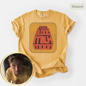May include: Mustard-colored t-shirt featuring a retro design with the text "HERMANOS GUTIERREZ BLOOD MILK MOON" in a bold, stylized font. The shirt has a classic crew neck and short sleeves. The shirt is made of a soft, comfortable material.