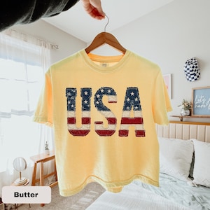 May include: A pale yellow short-sleeve t-shirt featuring the letters USA in a distressed American flag pattern. The letters are appliqued with a frayed edge finish. The shirt has a crew neckline and is displayed on a wooden hanger.