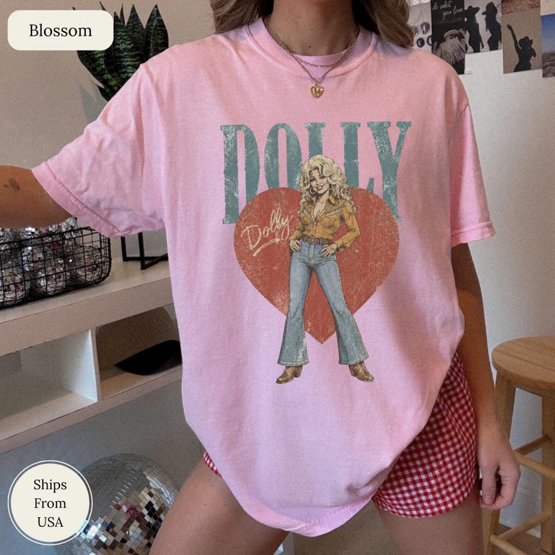 K&ouml;nnte beinhalten: Ein hellrosa kurz&auml;rmeliges T-Shirt mit einem gro&szlig;en roten Herzmotiv in Used-Optik. Im Herz befindet sich die Illustration einer Person mit blonden Haaren, die ein beiges Hemd und eine blaue Jeans tr&auml;gt. Das Wort DOLLY ist in gro&szlig;en blaugr&uuml;nen Buchstaben &uuml;ber dem Herz gedruckt, mit dem Namen Dolly in Schreibschrift auf dem Herz.
