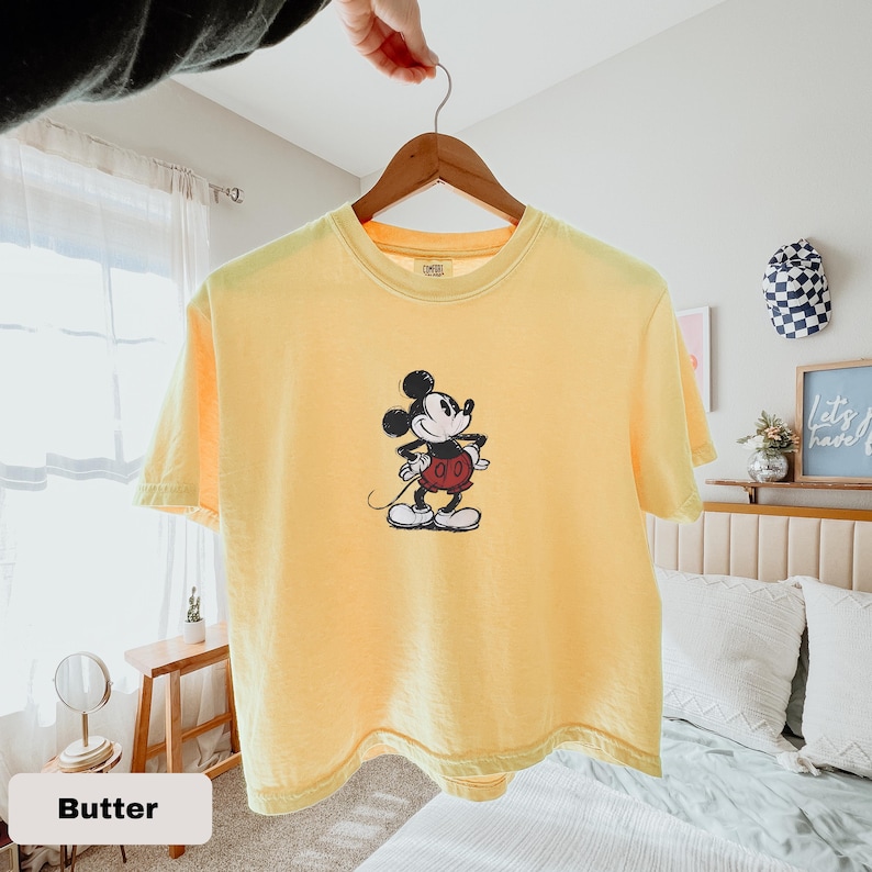 May include: A pale yellow t-shirt with a Mickey Mouse design. The cartoon character is in black, white, and red. The shirt is suspended from a wooden hanger.