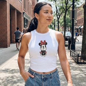 May include: White crop top featuring a Minnie Mouse graphic. The sleeveless top has a round neckline and a cartoon image of Minnie Mouse with a red bow. The top is worn with blue denim jeans.