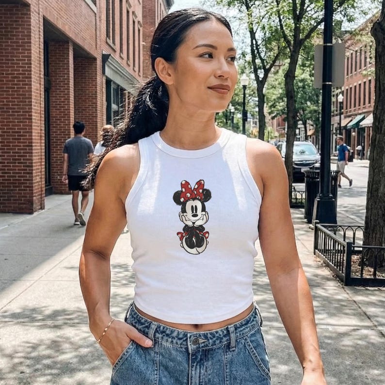 May include: White crop top featuring a Minnie Mouse graphic. The sleeveless top has a round neckline and a cartoon image of Minnie Mouse with a red bow. The top is worn with blue denim jeans.