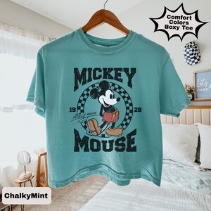 May include: A teal Comfort Colors boxy tee featuring a graphic of Mickey Mouse. The design includes the word "MICKEY" above a checkered circle with Mickey Mouse, the numbers "19" and "28", and the word "MOUSE" below.