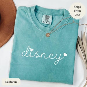 Puede incluir: Camiseta verde espuma de mar con la palabra "disney" en escritura blanca, con una cabeza de Mickey Mouse y un coraz&oacute;n. Un collar dorado con un colgante descansa sobre la camiseta. La camiseta est&aacute; etiquetada como "Comfort Colors" y es talla S-P-CH.