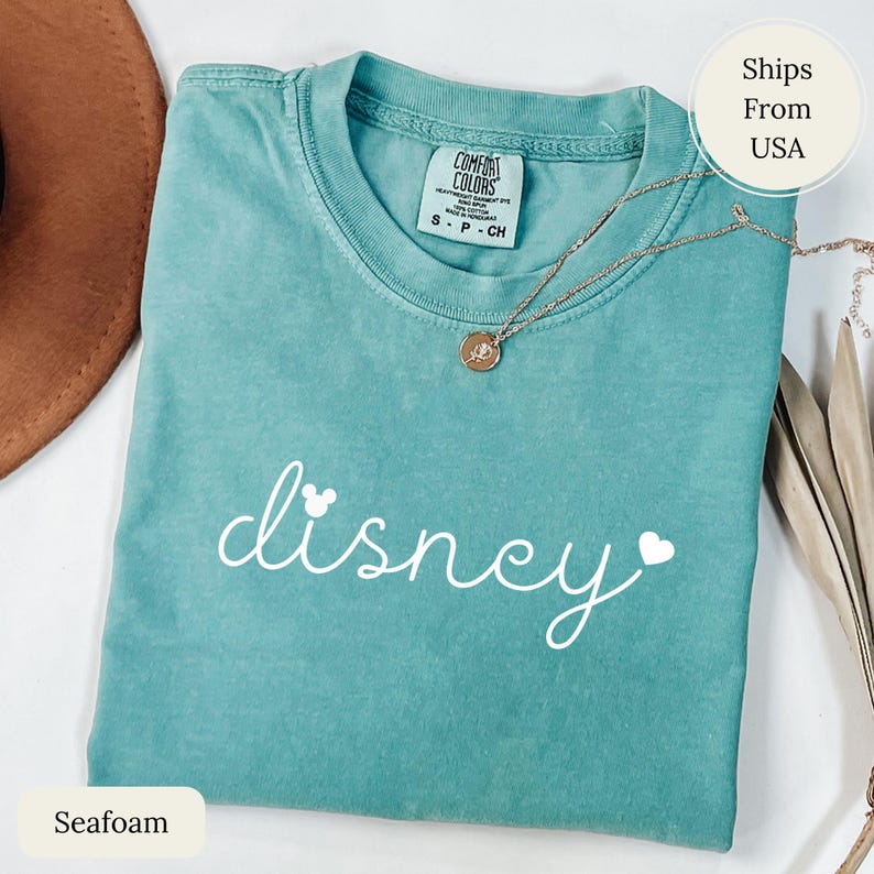 Puede incluir: Camiseta verde espuma de mar con la palabra "disney" en escritura blanca, con una cabeza de Mickey Mouse y un coraz&oacute;n. Un collar dorado con un colgante descansa sobre la camiseta. La camiseta est&aacute; etiquetada como "Comfort Colors" y es talla S-P-CH.