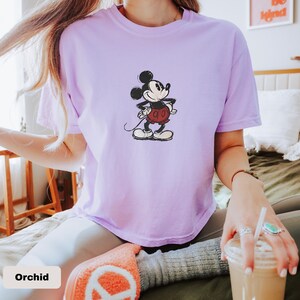May include: Orchid-coloured t-shirt featuring a classic Mickey Mouse graphic. The cartoon character is depicted in black, red, and white, with his hands on his hips. The shirt is a crew neck style.