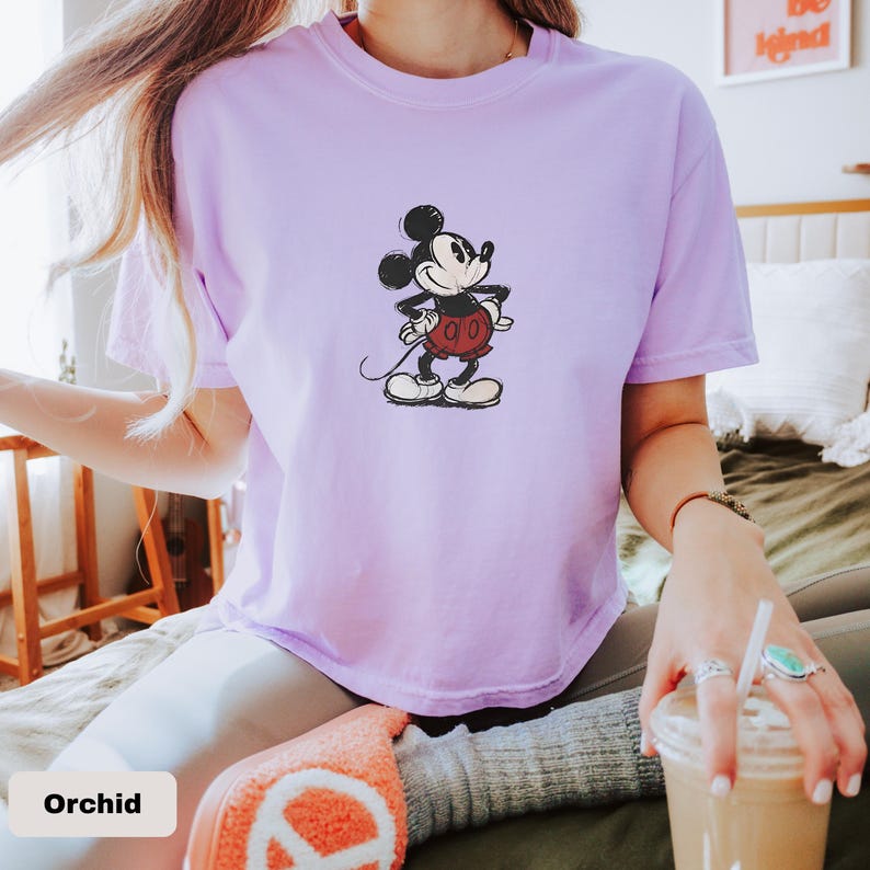 May include: Orchid-coloured t-shirt featuring a classic Mickey Mouse graphic. The cartoon character is depicted in black, red, and white, with his hands on his hips. The shirt is a crew neck style.