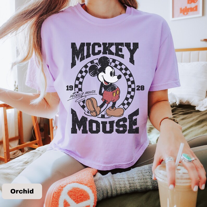 May include: Orchid-colored t-shirt featuring a distressed graphic of Mickey Mouse. The design includes the words "Mickey Mouse" and the years "1928" and "1928". The shirt has a classic crew neck and short sleeves.