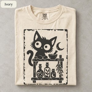May include: An ivory t-shirt featuring a black graphic print of a fluffy cat sitting on a wooden shelf with a mortar and pestle, a crescent moon, and three glass bottles containing liquid and herbs, all enclosed within a distressed rectangular border.