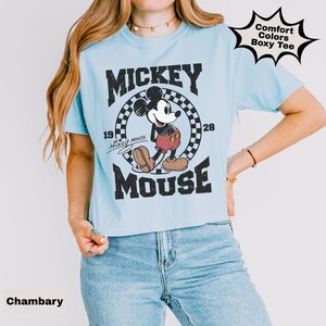 May include: Light blue t-shirt featuring a vintage-style graphic of Mickey Mouse. The design includes the words "Mickey Mouse" and the years "1928" and "19". The shirt also has the text "Comfort Colors Boxy Tee" in a starburst shape.