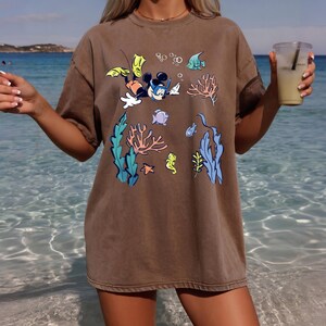 May include: Brown t-shirt featuring a cartoon Mickey Mouse snorkeling underwater with colorful coral, fish, and seaweed. The design includes bubbles and a seahorse. The shirt has a relaxed fit.