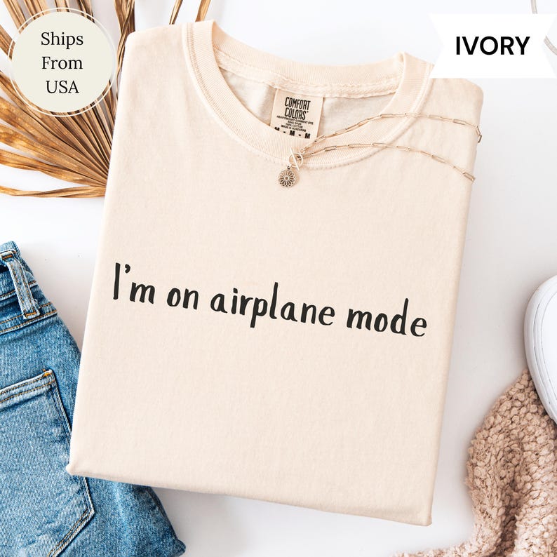 May include: An ivory-coloured t-shirt with the text "I'm on airplane mode" printed in black. The shirt has a crew neck and short sleeves. A gold necklace with a pendant is draped across the shirt. The shirt is folded on a white surface.