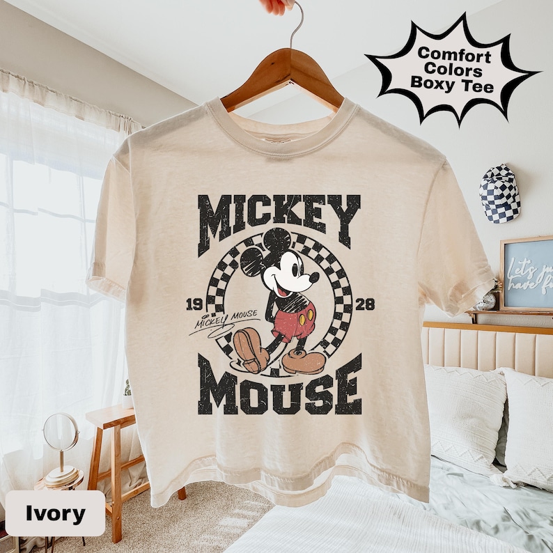 May include: Ivory-colored boxy tee featuring a vintage Mickey Mouse graphic. The design includes the words "Mickey Mouse" and the years "1928" and "19". The shirt is hanging on a wooden hanger.