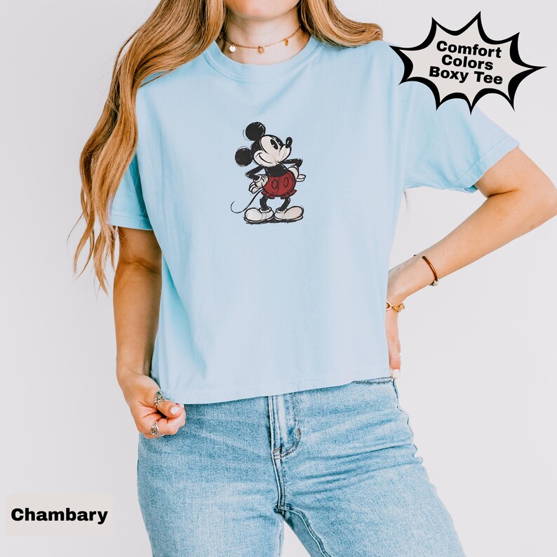 May include: Pale blue Comfort Colors Boxy Tee with a sketched Mickey Mouse design. The shirt has short sleeves and a crew neck. The word "Chambary" is printed at the bottom left.
