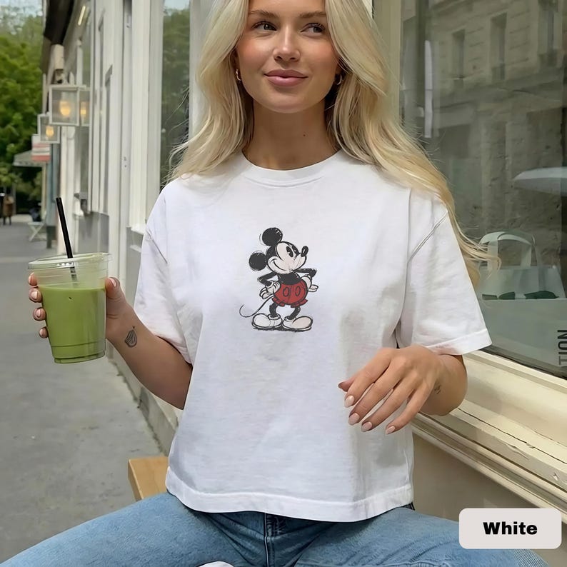 May include: White t-shirt with a Mickey Mouse graphic. The cartoon character is in black, red, and white. The shirt is a casual style, suitable for everyday wear.