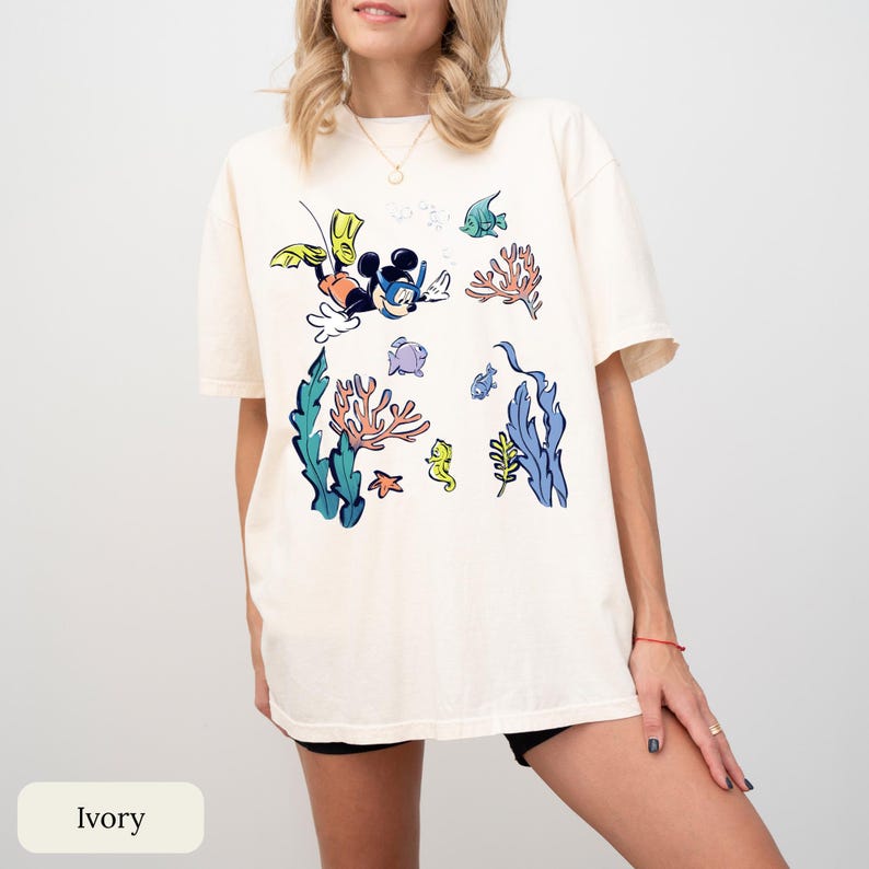 May include: Ivory-colored t-shirt featuring a cartoon graphic of Mickey Mouse scuba diving underwater. The design includes colorful coral, fish, and seaweed. The shirt is a relaxed fit.