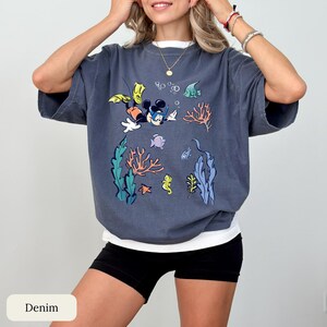 May include: A denim blue t-shirt featuring a colorful cartoon graphic of Mickey Mouse snorkeling underwater, surrounded by coral, fish, and seaweed. The shirt has a white undershirt and is paired with black shorts.