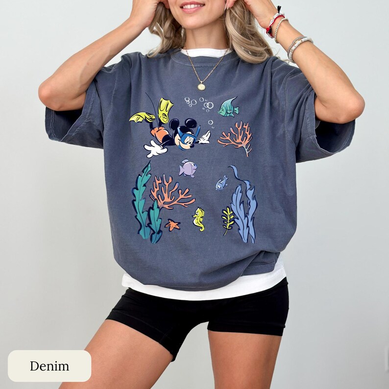 May include: A denim blue t-shirt featuring a colorful cartoon graphic of Mickey Mouse snorkeling underwater, surrounded by coral, fish, and seaweed. The shirt has a white undershirt and is paired with black shorts.