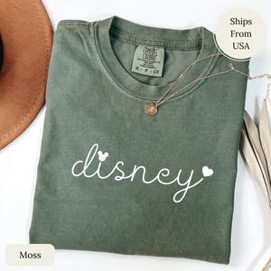 Puede incluir: Una camiseta verde musgo con la palabra "disney" en escritura blanca, con una cabeza de Mickey Mouse y un coraz&oacute;n. Un collar dorado con un colgante descansa sobre la camiseta. La camiseta es de material Comfort Colors.