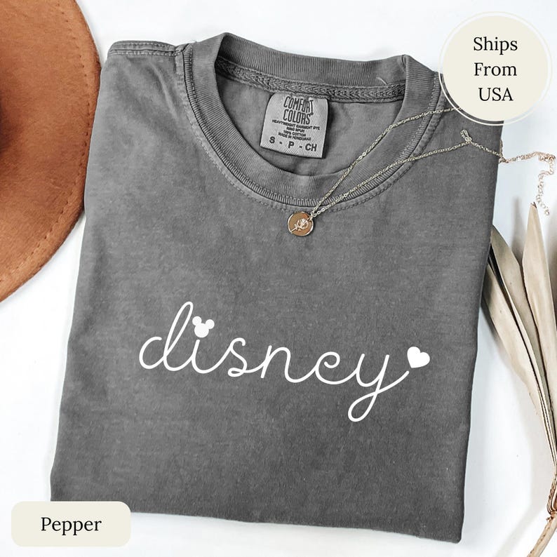 Puede incluir: Una camiseta gris Comfort Colors con la palabra "disney" en escritura blanca, con una cabeza de Mickey Mouse y un coraz&oacute;n. Un collar plateado con un colgante est&aacute; sobre la camiseta. La camiseta est&aacute; doblada y tiene una etiqueta que dice "Pepper".