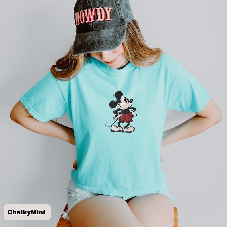 May include: A turquoise t-shirt with a Mickey Mouse design. The cartoon character is in black, red, and white. The person in the image is wearing a black baseball cap with the word "HOWDY" embroidered on it.