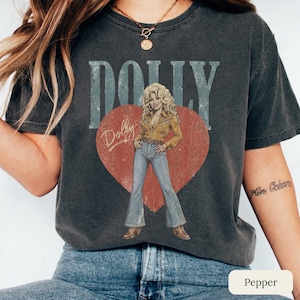 K&ouml;nnte beinhalten: Ein anthrazitfarbenes Kurzarm-T-Shirt mit einem gro&szlig;en roten Herz-Motiv, das eine blonde Person in einem gelben Hemd und blauer Schlaghose zeigt. Der Schriftzug DOLLY erscheint in gro&szlig;en hellblauen Buchstaben &uuml;ber dem Herz-Motiv.