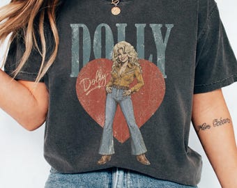 Puede incluir: Una camiseta gris carbón de manga corta con un gráfico de un gran corazón rojo que muestra a una persona rubia con camisa amarilla y pantalones azules de campana. El texto DOLLY aparece en letras grandes de color azul claro sobre el corazón.
