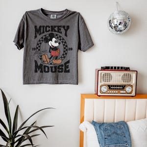 May include: Gray t-shirt featuring Mickey Mouse. The design includes the words "Mickey Mouse" and the years "1928" and "1928". The shirt is hanging on a white wall. A disco ball and a vintage radio are also visible.