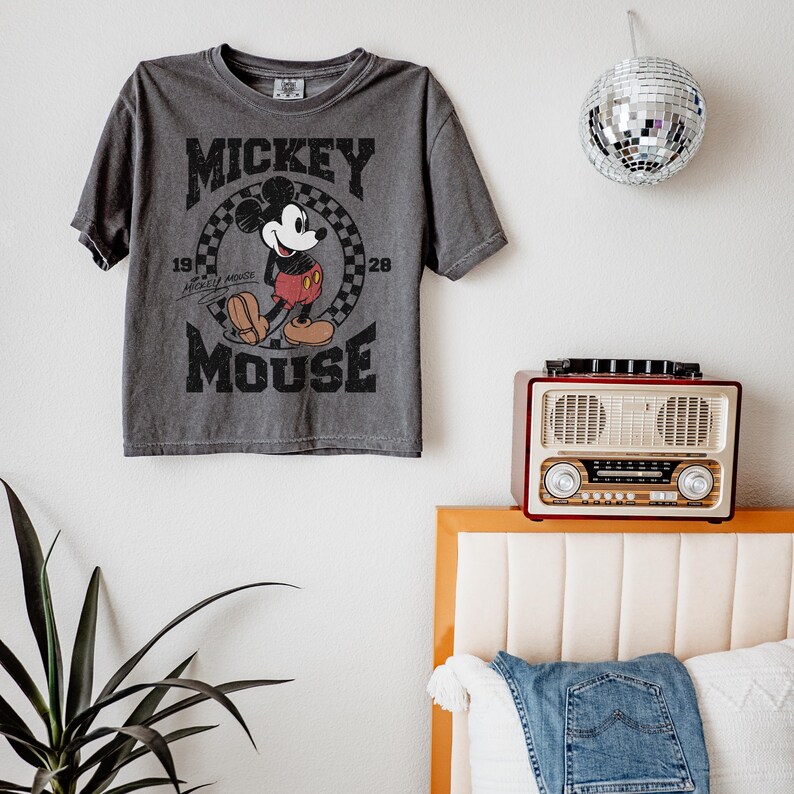 May include: Gray t-shirt featuring Mickey Mouse. The design includes the words "Mickey Mouse" and the years "1928" and "1928". The shirt is hanging on a white wall. A disco ball and a vintage radio are also visible.