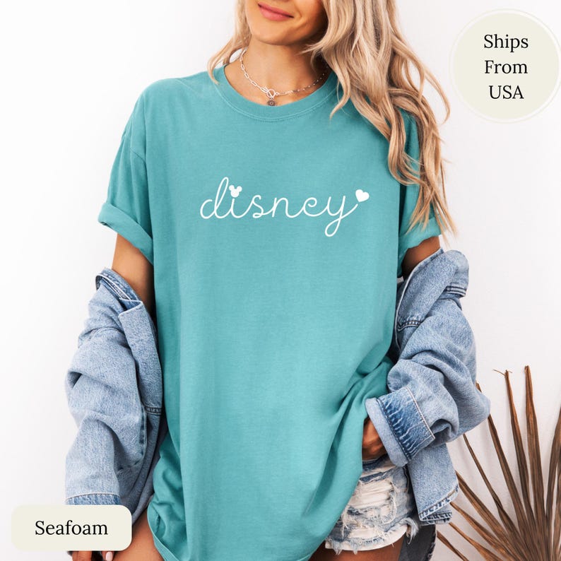 Puede incluir: Camiseta verde mar con la palabra "Disney" en escritura blanca. La palabra "Disney" tiene un coraz&oacute;n a cada lado. La camiseta es de corte holgado y tiene las mangas enrolladas.