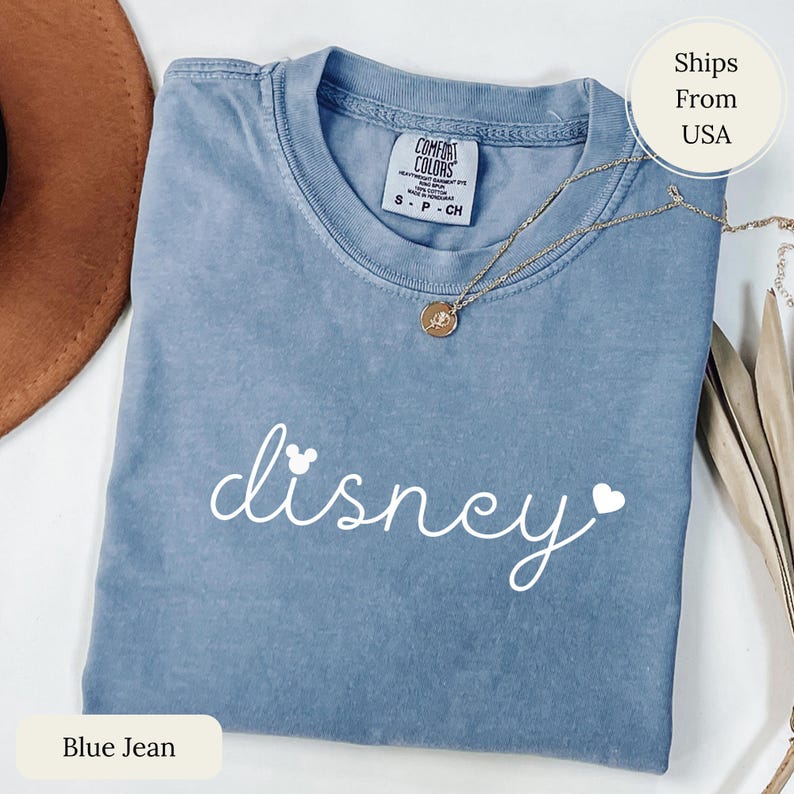 Puede incluir: Una camiseta azul vaquero con la palabra "disney" en escritura blanca, con una cabeza de Mickey Mouse y un coraz&oacute;n. Un collar de oro con un colgante descansa sobre la camisa. La camisa est&aacute; etiquetada como "Comfort Colors".