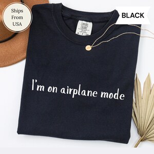 May include: Black crew neck t-shirt with the text "I'm on airplane mode" in white. A gold necklace with a small pendant is draped on the shirt. The shirt is folded and displayed on a white surface.