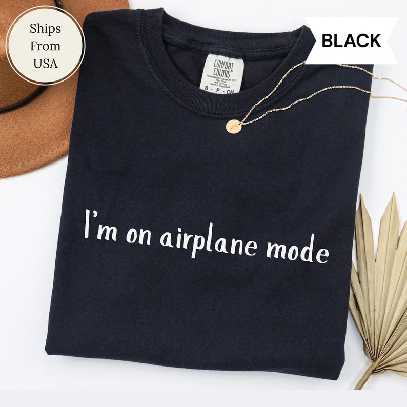May include: Black crew neck t-shirt with the text "I'm on airplane mode" in white. A gold necklace with a small pendant is draped on the shirt. The shirt is folded and displayed on a white surface.