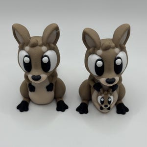 Puede incluir: Dos figuras de canguros marrones con grandes ojos con bordes blancos y detalles negros. Un canguro tiene una cría en su bolsa. Las figuras están sobre una superficie blanca.